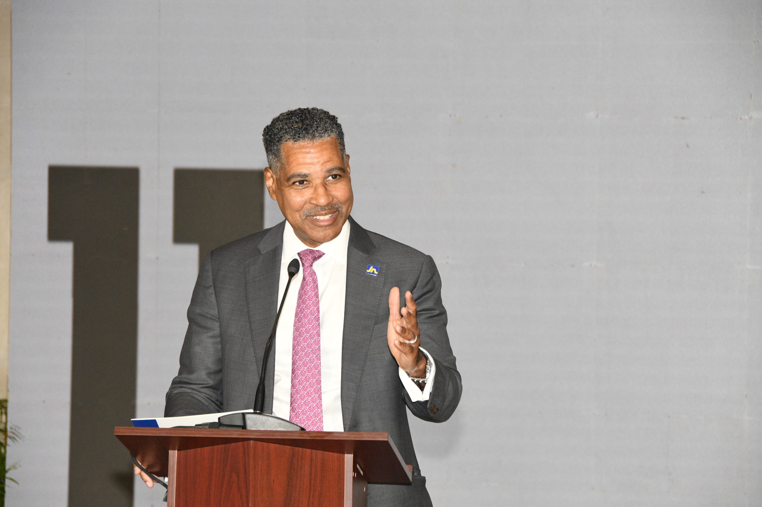 Keith Levy 5 - JN Bank Man in a gray suit and pink tie speaks at a wooden podium with a microphone, gesturing with his right hand at a formal event.