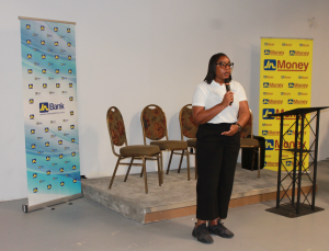 Monique - BramptonxJamaica - JN Bank Woman in a white top and black pants speaks into a microphone on a small stage, with JA Bank and Money banners nearby.