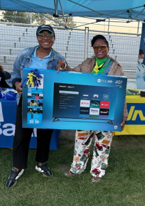 Monique and JN Member - JN Bank Two women under a blue canopy hold a boxed 40-inch Philips Roku TV prize at an outdoor event.