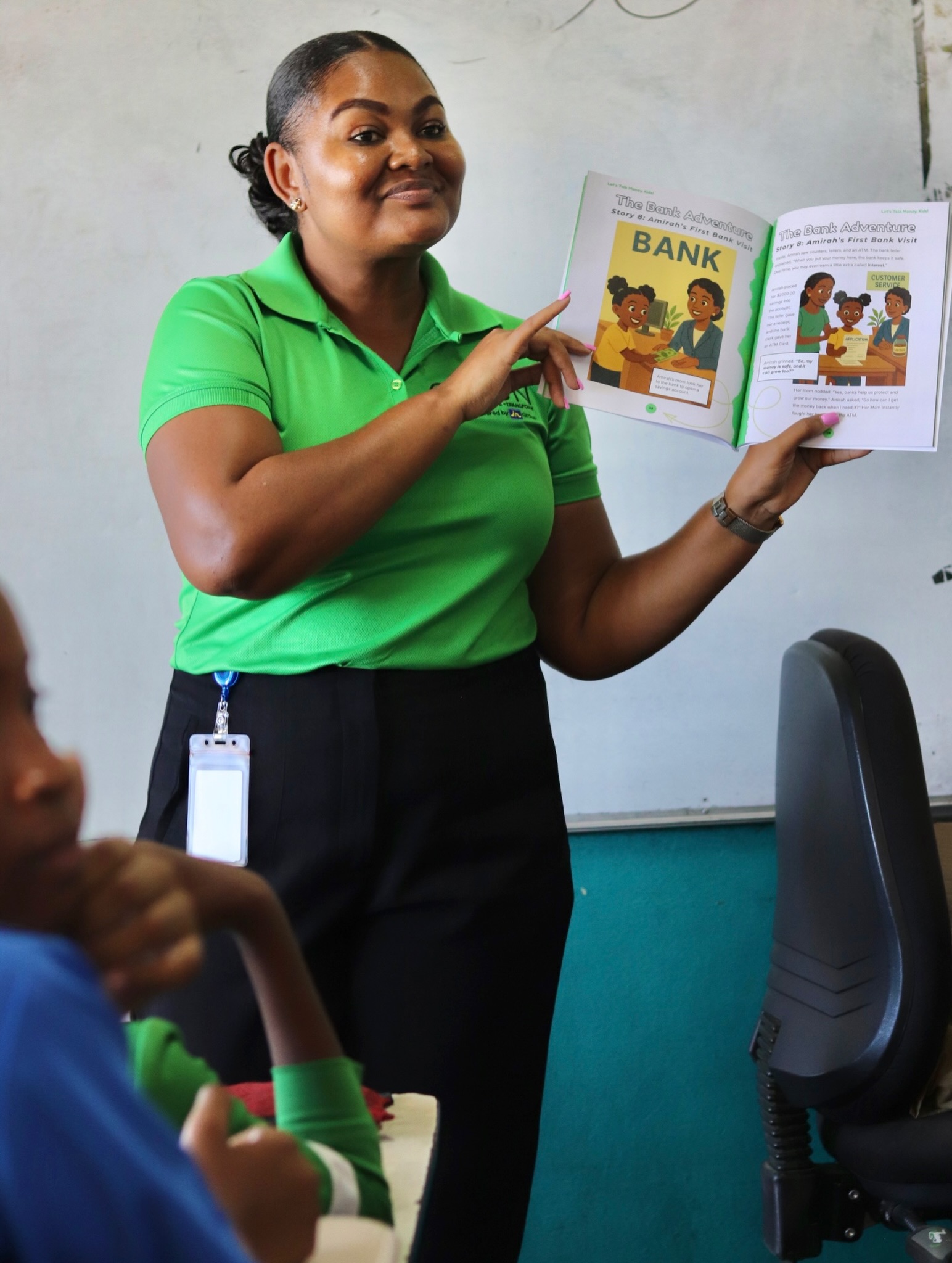 Tomoya Swaby reading - JN Bank Woman in a bright green polo holds up an illustrated children's book about a bank, presenting to students in a classroom.