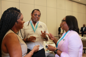 20251028_154551780_iOS - JN Bank Three professionals converse at a conference, with two women and a man wearing name badges and teal lanyards, standing by a table with materials.