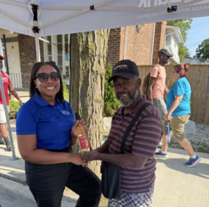 Image - Farmworker - JN Bank Woman in a blue polo hands a bottled drink to a man in a striped polo under a white canopy at a community event; both smiling, with other volunteers walking in the background.